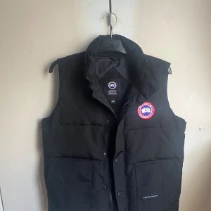 Svart dunväst från Canada Goose - Svart dunväst från Canada Goose med hög krage och klassisk logga på bröstet. Västen har två stora fickor framtill och stängs med både dragkedja och knappar. Perfekt för lager-på-lager och riktigt snygg till streetwear.