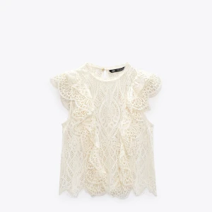 Vit spetsblus med volang från Zara - Superfin vit blus från Zara i spets med snygga volangdetaljer framtill och på axlarna. Ärmlös modell med hög hals och vackert mönstrad spets som ger en romantisk vibe. Perfekt att styla med jeans eller kjol för en trendig look.