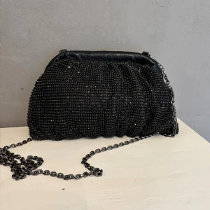 Svart glittrig clutch med kedja - Elegant svart kuvertväska med glittrande detaljer och en stilren kedja som axelrem. Köpt för 569 kr. Liten defekt på kedjan vilket syns på sista bilden. Annars är väskan i väldigt bra skick. Längden är ca 23 cm, höjden är ca 15 cm och kedjan är ca 114 cm totalt. Märket är mascara london.