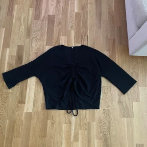 Svart croppad topp med snörning - Svart croppad topp med v-ringning och snörning framtill som ger en snygg rynkad effekt. Toppen har trekvartsärm och är tillverkad i ett ribbat material som känns mjukt mot huden. Perfekt för dig som gillar stilrena och trendiga plagg.