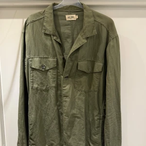 Olivgrön overshirt  - Snygg olivgrön overshirt från East West med klassisk krage och två stora bröstfickor med lock och knapp. Skjortan har knäppning framtill och är tillverkad i ett mjukt bomullstyg. Perfekt för lager-på-lager och chill stil. XL