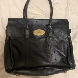 Svart Mulberry Bayswater handväska  - Säljer en klassisk svart Mulberry Bayswater handväska med strukturerad yta. Väskan har guldiga metalldetaljer, ikonisk vridknapp och dubbla handtag. Insidan har dragkedjefack och Mulberrys logga i guld. Perfekt för dig som gillar lyxiga accessoarer.