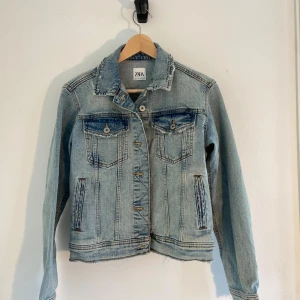 Ljusblå jeansjacka från Zara - Snygg ljusblå jeansjacka från Zara med klassisk krage, knappar framtill och två bröstfickor med lock. Jackan har slitna detaljer vid kragen och nederkanten för en cool, avslappnad look. Perfekt för dig som gillar streetstyle och denim.