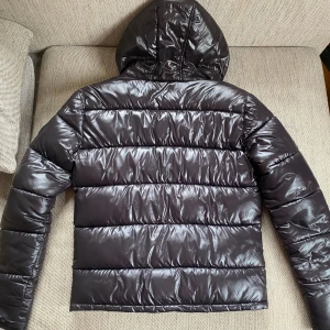 Svart pufferjacka Supply & Demand XS - Snygg svart pufferjacka från Supply & Demand med glansig finish, huva med broderad logga och två dragkedjefickor framtill. Jackan har quiltad design och patch med logga på ärmen. Perfekt för kalla dagar och streetwear-stil.