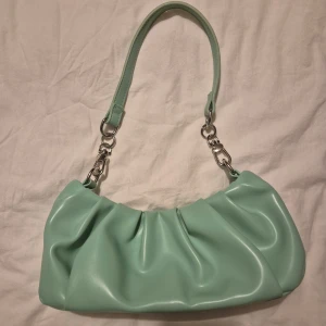 Mintgrön axelväska i skinnimitation - Superfin mintgrön axelväska med snygga veck och silverfärgade metalldetaljer. Väskan har avtagbar axelrem och stängs med magnetknapp. Inuti finns ett svart foder och en innerficka med dragkedja. Perfekt för att lyfta din outfit med en pop av färg.