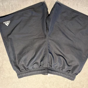 Svarta badbyxor från Adidas - Svarta träningsshorts från Adidas med klassisk logga på ena benet. Shortsen har elastisk midja och är tillverkade i ett lätt och ventilerande syntetmaterial, perfekta för sport och träning. Enkel och stilren design som funkar till alla sportiga outfits.