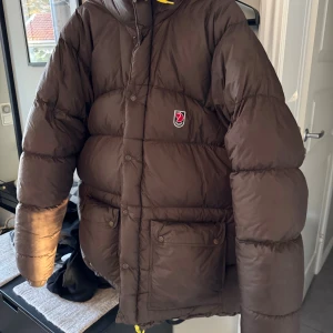 Fjällräven Expedition XL - Säljer en brun pufferjacka från Fjällräven i storlek XL. Jackan har huva med gula snören, stora fickor med knappar och grov dragkedja. Perfekt för kalla dagar och har en schysst, oversized look. Klassisk Fjällräven-logga på bröstet.