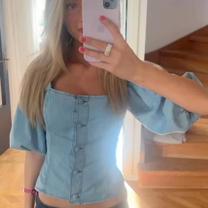 Ljusblå korsettopp med puffärm - Superfin ljusblå korsettopp i denim-look med puffiga korta ärmar och knappar framtill. Toppen har fyrkantig urringning och markerad midja som ger en snygg siluett. Perfekt för dig som vill sticka ut med en trendig och cool stil💕