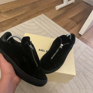 Svarta sneakers Axel Arigato mocka - Snygga svarta sneakers från Axel Arigato i mocka med dragkedja framtill istället för klassisk snörning. Skorna har rund tå, platt sula och detaljer i svart läder vid hälen. Perfekta för dig som gillar stilrena och moderna skor.box medföljer ej 