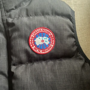 Svart dunväst från Canada Goose M - Svart dunväst från Canada Goose med klassisk logga på bröstet och ståkrage. Västen har quiltad design, tryckknappar framtill och två fickor med knappstängning. Perfekt för kalla dagar och snygg att styla över hoodie eller tröja.