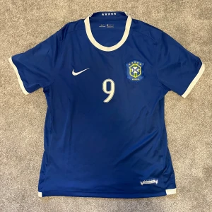 Blå Brasil Ronaldo 9 Nike fotbollströja - Säljer en blå Brasilien fotbollströja från Nike med Ronaldo och nummer 9 på ryggen. Tröjan har vita detaljer vid krage och ärmslut, samt Brasilien-emblem på bröstet. Tillverkad i lätt och ventilerande material, perfekt för fotboll eller samling.