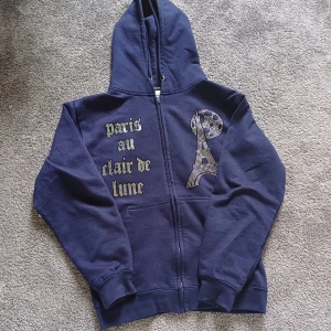 Mörkblå bling lucid zip hoodie med strass och tryck - Mörkblå hoodie från BLINGLUCID med dragkedja, huva och fickor. Framsidan har texten 'paris au clair de lune' och ett motiv av Eiffeltornet och månen i strass. Tillverkad i mjuk bomull och polyester, perfekt för dig som gillar unika detaljer.