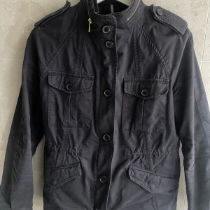 Svart parkajacka från J.Lindeberg - Svart parkajacka från J.Lindeberg med snygga axelklaffar, fyra fickor framtill och markerad midja. Jackan har både knappar och dragkedja framtill samt hög krage. Perfekt för dig som vill ha en stilren och funktionell jacka.