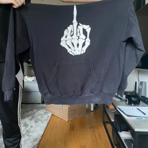 Svart hoodie med skelett-tryck - Svart hoodie med stor ficka framtill och huva. På bröstet finns ett vitt tryck av en skelett-hand som visar långfingret. Perfekt för dig som gillar edgy och unika plagg. Klassisk passform och mjukt material. Storlek L men skulle säga att den snarare är som en S