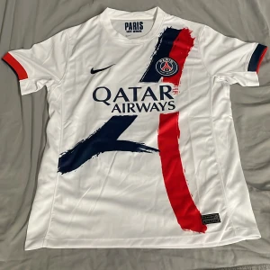 PSG vit kortärmad fotbollströja Nike - Paris Saint-Germain matchtröja i vitt med coola penseldrag i marinblått och rött på framsidan. PSG-logga på bröstet, Qatar Airways tryck och Nike Swoosh. Tillverkad i lätt Dri-Fit polyester som andas. Röd och marinblå detalj på ärmslut.