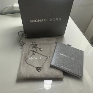 Silver halsband med hjärta från Michael Kors - Snyggt silverfärgat halsband från Michael Kors med ett hjärtformat hänge dekorerat med små glittriga stenar och MK-logga i mitten. Kedjan är tunn och elegant, perfekt för att ge din outfit en lyxig touch. Levereras med originalpåse och ask. Nypris 1500
