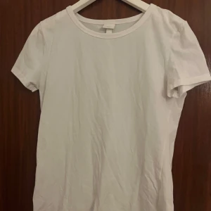 Vit basic t-shirt från H&M - En klassisk vit t-shirt från H&M Basics i storlek M. Modellen har rund halsringning och korta ärmar, tillverkad i mjuk bomull som är skön mot huden. Perfekt att matcha med jeans eller shorts för en enkel och clean look.