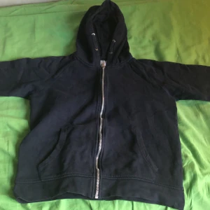 Svart hoodie med dragkedja från 157 - Svart hoodie från 157 med hel dragkedja framtill och stor huva. Tröjan har känguruficka och är tillverkad i mjuk bomullsblandning. Perfekt för chill dagar eller när du vill ha en enkel och stilren look.