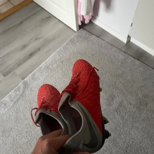 Röda Nike fotbollsskor med dobbar - Säljer ett par röda Nike fotbollsskor med Flyknit ACC-teknologi. Skorna har snörning, silvergrå detaljer och robusta metall-dobbar för gräsplan. Ovandelen har ett mönstrat, hexagonformat struktur för extra grepp och kontroll. Perfekt för dig som vill ha snygga och funktionella skor på planen. Liten defekt finns på sista bilden!