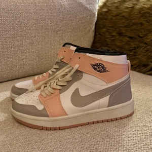 Nike Air Jordan 1 High  - Nike Air Jordan 1 High sneakers i en snygg mix av ljusrosa, grå och vit. Skorna har klassisk snörning, rund tå och högt skaft. Ikoniska Air Jordan-loggan på sidan och bak, med mjuk vadderad insida och slitstark yttersula. Perfekt för dig som vill sticka ut med stil.