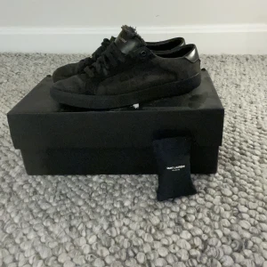 Svarta sneakers från Saint Laurent - Svarta Saint Laurent Paris sneakers med snyggt fransad kant vid plösen och ton-i-ton logga. Skorna har rund tå, platt sula och snörning. Materialet är mocka med detaljer i skinn, vilket ger en lyxig och cool vibe. Perfekta för dig som gillar stilrena och exklusiva sneakers. Pris går att diskutera. [ skick 7,5 av 10] snöre är tyvär lite sönder följer med påse till skosnöre och box är storlek för 40 akter som 41. Bara att höra av sig vid frågor ☺️ dom är äkta