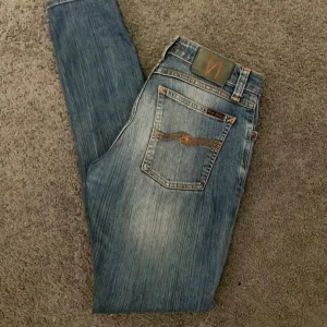 Nudie Jeans blå straight fit jeans - Säljer ett par klassiska blå jeans från Nudie Jeans med orange sömmar och ikonisk brodyr på bakfickan. Jeansen har normal passform och raka ben, tillverkade i slitstarkt bomullstyg. Perfekta för dig som gillar en tidlös och avslappnad look. Storleken är 29/32 