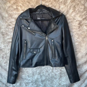 Svart skinnjacka från Zara - Snygg svart skinnjacka från Zara med klassisk bikerstil. Jackan har flera silvriga dragkedjor, bred krage med tryckknappar och detaljerade fickor. 