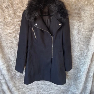 Svart kappa med fuskpäls från New Look M - Svart kappa från New Look med stor fluffig fuskpäls på kragen och snedställd dragkedja. Slits baktill. Storlek 10 motsvarar M.