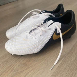 Nike Phantom fotbollsskor vit/guld - Nike Phantom fotbollsskor i vitt med svarta och guldiga detaljer. Snygg design med guldigt swoosh på sidan och mönstrad ovandel. Skorna har snörning och är tillverkade i syntetmaterial för bästa grepp på planen.