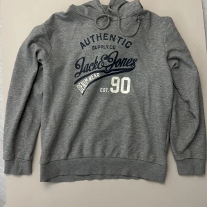 Grå hoodie från Jack & Jones - Snygg grå hoodie från Jack & Jones med tryck framtill i blått och vitt. Tröjan har huva med snörning och är tillverkad i mjuk bomullsblandning. Perfekt för chill dagar eller när du vill ha en avslappnad stil.