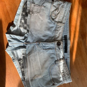 Ljusblå denimshorts från ONLY - Säljer ett par ljusblå denimshorts från ONLY med slitna detaljer framtill och klassiska fem fickor. Shortsen har låg midja och coola stjärnor på fickfodret. Perfekta för varma dagar och en avslappnad stil.
