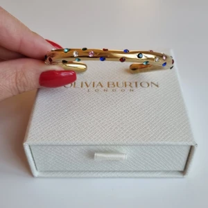 Guldfärgat armband  - Stilrent guldfärgat armban med rundad form och dekorerat med små färgglada stenar i blått, rött, grönt, rosa och vitt. Perfekt statement-piece för dig som gillar färg och glittriga detaljer. Lådan ingår ej.