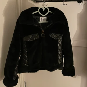 Svart pälsjacka från Jayriss med quiltade detaljer - Säljer en svart pälsjacka från Jayriss med snygga quiltade detaljer i skinnimitation på fickor och sidor. Jackan har en stor krage, dragkedja med ring och långa ärmar. Perfekt för dig som vill sticka ut med en cool och mysig look. Passar både i hösten🍁och i vintern❄️. Är size L men funkar M.