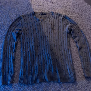 Grå kabelstickad tröja från Polo Ralph Lauren - Säljer en klassisk grå kabelstickad tröja från Polo Ralph Lauren med rund halsringning och broderad logga på bröstet. Tröjan har långa ärmar och ribbade muddar vid ärmslut och nederkant. Perfekt för lager-på-lager-stil.