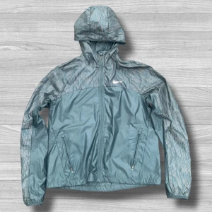 Nike vindjacka med huva, blå M - Nike vindjacka i blått med reflekterande detaljer och mönstrade partier upptill. Jackan har huva, dragkedja framtill och två fickor med dragkedja. Perfekt för löpning eller utomhusaktiviteter. Lätt och smidig modell med sportig look.