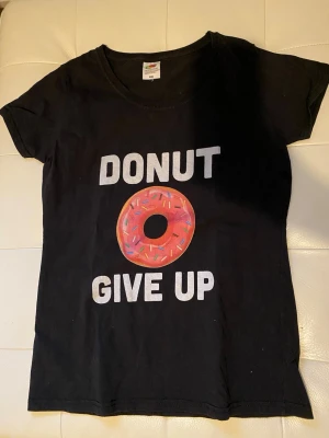 Svart t-shirt med donut-tryck - Svart t-shirt från Fruit of the Loom i modellen Lady Fit Valueweight, storlek S. Framsidan har ett stort tryck med texten 'DONUT GIVE UP' och en färgglad donut med strössel. T-shirten har rund hals och korta ärmar, perfekt för dig som gillar roliga prints.