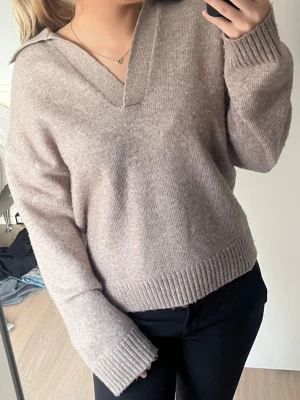 Beige stickad tröja med v-ringning - Säljer en beige stickad tröja med bred v-ringning och krage. Tröjan har långa ärmar och ribbade muddar vid ärmslut och nederkant. Perfekt för dig som gillar en chill och stilren look.