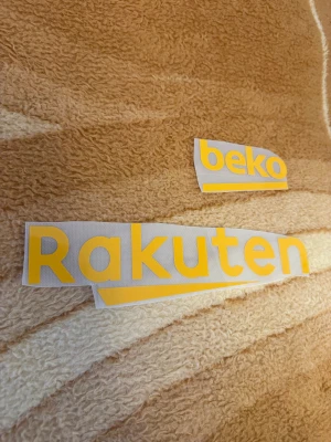 Fc Barcelona sponsor tryck - Säljer en fc Barcelona tryck med texten 'beko' och 'Rakuten' i gul färg på vit bakgrund.