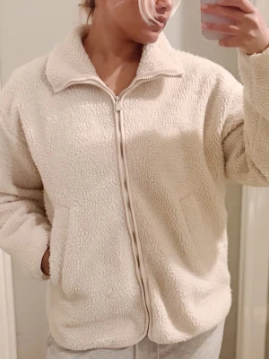 Vit/Beige teddyjacka från McKINLEY - Mysig beige teddyjacka med dragkedja framtill och två fickor. Jackan har en hög krage och är tillverkad i fluffigt fleece-material. 