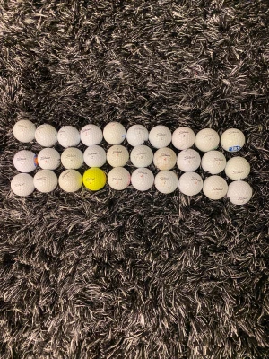 30 titeliest bollar A till F klass - Bilden innehåller ett antal golfbollar av märket Titleist, varav en är gul och resten vita. Golfbollarna är placerade på en mörk, lurvig matta. Ingen sko syns på bilden.