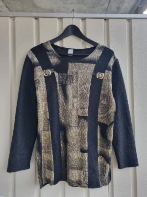 Svart och guldig långärmad tröja  - Unik långärmad tröja med  två dekorativa spännen framtill. Materialet är antagligen stickat. Köpt på second hand och används ett få tal gånger. Svårt att tyda vilket märkte det är pga suddig lapp, se bild.