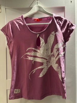 Lila t-shirt med tryck från Puma - Säljer en lila t-shirt från Puma med ett stort vitt blommigt tryck på framsidan. T-shirten har rund hals med en liten öppning och korta ärmar. Materialet är mjuk bomull som känns skönt mot huden. Perfekt för dig som gillar sportiga och färgstarka plagg.