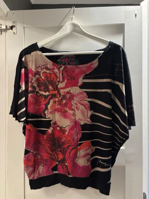 Blommig topp från Desigual - Säljer en snygg topp från Desigual med stora blommor i rött och rosa på svart botten. Toppen har bred halsringning, korta ärmar och ett coolt randigt mönster i beige. Materialet är mjukt och lätt, perfekt för att sticka ut med färg och mönster.