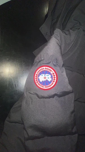 Svart ufferjacka XS - Svart pufferjacka från orlek XS. Jackan har huva, synliga knappar framtill och det klassiska röda Canada Goose-märket på ärmen. Fylld med dun och har en clean, stilren look som funkar när det är kallt ute.