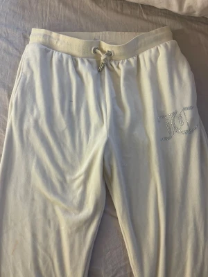 Vita mjukisbyxor från Juicy Couture - Säljer ett par vita mjukisbyxor från Juicy Couture med glittrande logga på ena benet. Byxorna har snörning i midjan och två bakfickor. Mjukt material som känns skönt mot huden, perfekt för chill dagar. Storlek 15/16.