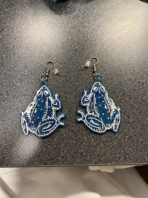 Blå glittriga grodörhängen - Unika örhängen i form av grodor, gjorda i blått glittrigt material med vita detaljer på framsidan. Baksidan är helblå med inslag av glitter och små färgprickar. Perfekt för dig som vill sticka ut med coola accessoarer!