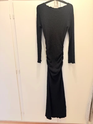 Svart långklänning från Gina Tricot - Säljer en svart långklänning från Gina Tricot i storlek S. Klänningen har ett ribbat, texturerat tyg och långärmade ärmar. Den är figurnära med snygg rynkning längs midjan och har en enkel, stilren design som passar perfekt för dig som gillar minimalistiskt mode.