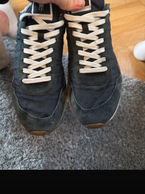 Svarta Polo sneakers med vit sula - Säljer ett par svarta Polo sneakers med vit sula och bruna gummisulor. Skorna har vit snörning och detaljer i mocka och syntet. Polo-logga syns både på hälen och under sulan. Klassisk rund tå och platt sula, perfekt för dig som gillar stilrena sneakers.