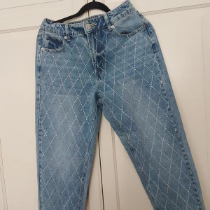 Blå jeans med strass från Amisu, stl 38 - Snygga ljusblå jeans från Amisu i storlek 38. Jeansen har raka ben och är dekorerade med ett rutmönster av strass framtill som ger en unik och glittrig look. Klassisk femficksmodell och normal passform. Perfekta för dig som vill sticka ut lite extra.