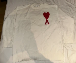 Vit stickad tröja från Ami Paris - Vit stickad tröja från Ami Paris med rund hals och ribbade muddar. På bröstet finns ett stort rött hjärta och bokstaven A som är typiskt för märket. Tröjan har en clean look och är perfekt för dig som gillar stilrena detaljer och vill sticka ut med en ikonisk design.
